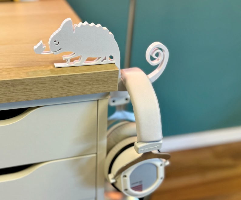 May include: White 3D printed chameleon headphone stand with a curled tail. The stand is mounted on a white desk with drawers. A pair of white headphones are hanging from the stand.