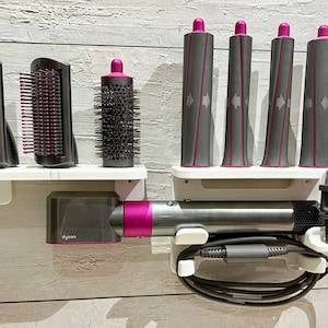 May include: A white wall-mounted organizer with a Dyson hair dryer, a round brush, two paddle brushes, and five curling iron attachments. The organizer has a white shelf and a black cord.