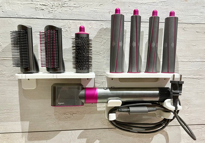 May include: A white wall-mounted organizer with a Dyson hair dryer, a round brush, two paddle brushes, and five curling iron attachments. The organizer has a white shelf and a black cord.