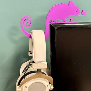 May include: Purple chameleon-shaped headphone stand mounted on a black computer monitor. The stand has a curled tail that hooks over the top of the monitor. A pair of white headphones are hanging on the stand.