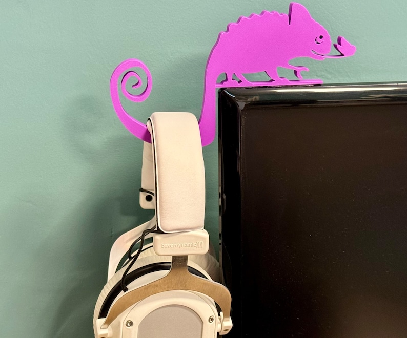 May include: Purple chameleon-shaped headphone stand mounted on a black computer monitor. The stand has a curled tail that hooks over the top of the monitor. A pair of white headphones are hanging on the stand.