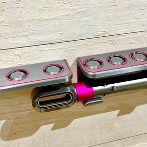May include: A gray wall-mounted storage rack with four circular slots for holding styling tools. A pink and silver hair styling tool is attached to the rack with the word "Dyson" printed on it.