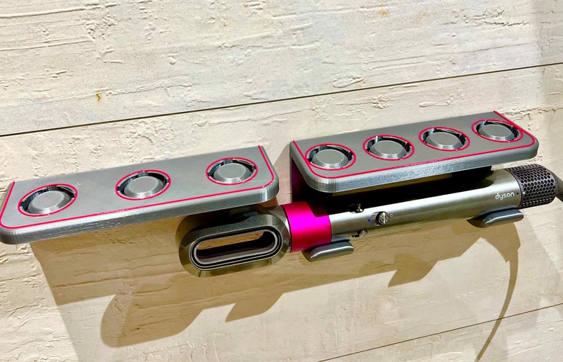 May include: A gray wall-mounted storage rack with four circular slots for holding styling tools. A pink and silver hair styling tool is attached to the rack with the word "Dyson" printed on it.