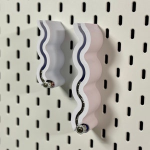 Könnte beinhalten: Zwei weiße Batteriehalter aus Kunststoff mit wellenförmigem Design. Die Halter sind an einer weißen Lochwand befestigt.