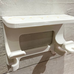 May include: White plastic toothbrush holder with four toothbrush slots and two hooks for hanging items. The holder is mounted on a wall.