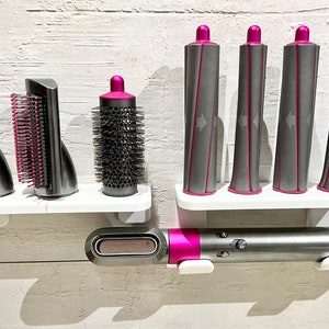 May include: A set of hair styling tools, including a curling iron, a round brush, and a comb attachment. The tools are mounted on a white wall-mounted shelf. The curling iron is pink and gray, and the other tools are gray.
