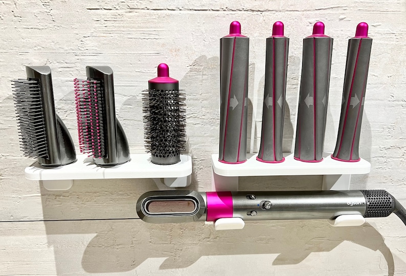May include: A set of hair styling tools, including a curling iron, a round brush, and a comb attachment. The tools are mounted on a white wall-mounted shelf. The curling iron is pink and gray, and the other tools are gray.