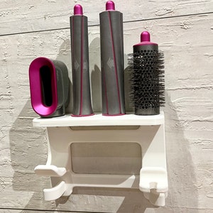 Può includere: Organizzatore da parete bianco per strumenti per lo styling dei capelli con quattro slot. Tre slot contengono arricciacapelli rosa e grigi e uno slot contiene una spazzola rotonda con setole nere.