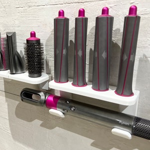 May include: A set of hair styling tools, including a Dyson Airwrap styler, a round brush, and four curling barrels. The tools are mounted on a white wall-mounted shelf. The curling barrels are gray with pink accents.