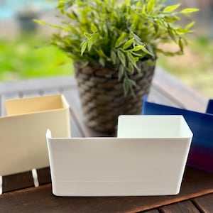 May include: Three plastic storage containers in different colors: beige, white, and blue. The containers are on a wooden table.