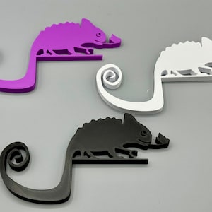 May include: Three chameleon-shaped phone holders in purple, white, and black. Each holder has a curled tail and a small leaf in its mouth.
