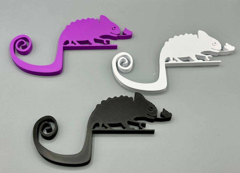 May include: Three chameleon-shaped phone holders in purple, white, and black. Each holder has a curled tail and a small leaf in its mouth.