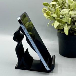 May include: A black phone stand shaped like a cat, holding a silver smartphone. The stand is on a white surface, with a potted green plant in the background.