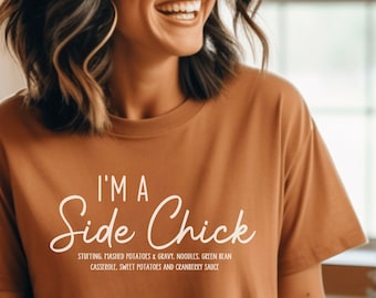 May include: A woman wearing a brown t-shirt with the text "I'm a Side Chick" and a definition of the term. The text is in white. The woman is smiling and looking to the right.