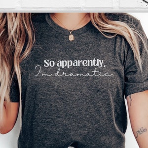 May include: A gray t-shirt with the text "So apparently, I'm dramatic" printed in white on the front. The shirt is being worn by a person with long blonde hair.