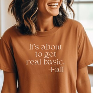 May include: A woman wearing a brown t-shirt with the text "It's about to get real basic. -Fall" printed on it.