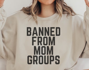 May include: A woman wearing a beige sweatshirt with the text "BANNED FROM MOM GROUPS" printed on it. She is wearing a white beanie and smiling. She is making the peace sign with both hands.