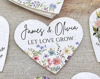 May include: Heart-shaped wedding favors with floral designs and the names "James & Olivia" and the phrase "LET LOVE GROW" printed on them. The favors feature various flower arrangements in shades of pink, blue, and yellow.