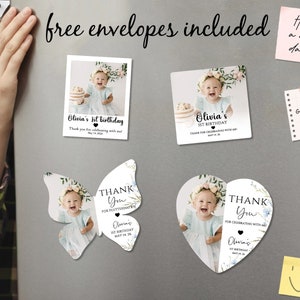 May include: A selection of personalized birthday and thank you cards, including butterfly and heart-shaped designs. The cards feature a baby's photo, floral accents, and the text "Olivia's 1st Birthday" and "Thank You". The phrase "free envelopes included" is also visible.