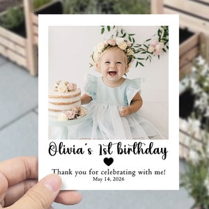 May include: A photo card featuring a baby girl in a light blue dress, celebrating her first birthday. The card says "Olivia's 1st birthday" with a heart and the text "Thank you for celebrating with me! May 14, 2026." The words "free envelopes included" are written above the card.