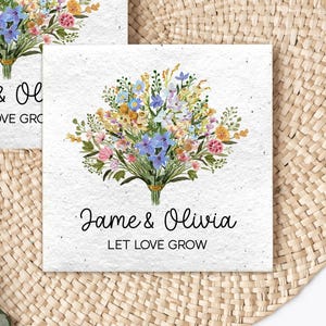 May include: Two white cards with floral bouquets and the text "Jame & Olivia" and "LET LOVE GROW". The bouquets feature blue, yellow, pink, and orange flowers. The cards are on a woven, circular placemat with green foliage.
