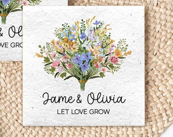 May include: Two white cards with floral bouquets and the text "Jame & Olivia" and "LET LOVE GROW". The bouquets feature blue, yellow, pink, and orange flowers. The cards are on a woven, circular placemat with green foliage.