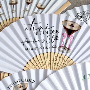 Può includere: Ventagli di carta bianca con un design a coste e manici in legno. I ventagli presentano testo e illustrazioni relative a una festa di compleanno, tra cui bicchieri da martini e la frase "A Tini Bit Older".