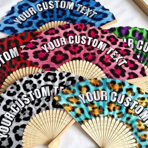 Op de afbeelding: Een verzameling van zes opvouwbare handwaaiers met houten ribben en stoffen hoezen met diverse luipaardprintpatronen in blauw, rood, roze, groen, grijs en geel. Elke waaier toont de witte tekst YOUR CUSTOM TEXT in het midden van de waaier.