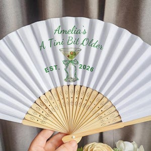 May include: White folding fan with a light brown wooden handle. The fan has a green martini glass graphic and the text "Amelia's A Tini Bit Older EST. 2026". The fan is open and held by a hand.