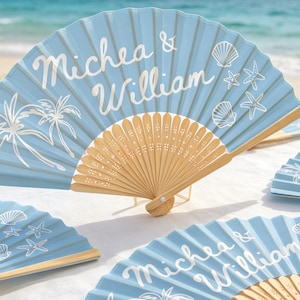 May include: A light blue folding hand fan with a light-colored wooden frame. The fan features white printed graphics of palm trees, seashells, and starfish, along with the white text Michea & William written in a cursive script font.