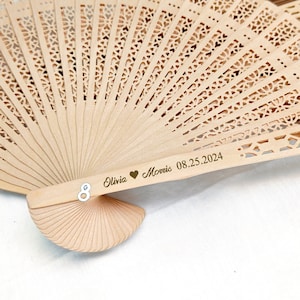 May include: A set of wooden hand fans with a delicate, intricate design. The fans are beige and have a personalized inscription that reads "Olivia & Morris 08.25.2024".
