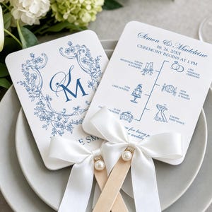 May include: Two white cardstock wedding programmes with rounded corners, featuring blue floral monograms and a timeline of events for Simon and Madeleine. Each card is attached to a wooden handle and decorated with a white satin ribbon and a pearl.
