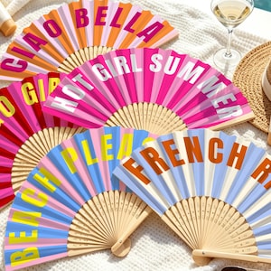 Può includere: Una collezione di quattro ventagli pieghevoli con stecche in legno e pannelli in tessuto. I ventagli presentano colori vivaci e scritte in grassetto tra cui CIAO BELLA, HOT GIRL SUMMER, LET'S GO GIRLS, BEACH PLEASE e FRENCH RIVIERA.