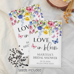May include: A white card with a floral design and the text "LOVE in bloom" with butterflies. The card also includes the text "MARISSA'S BRIDAL SHOWER 09.23.2024 WILDFLOWER SEEDS". A small pile of black seeds is in the lower left corner of the image.