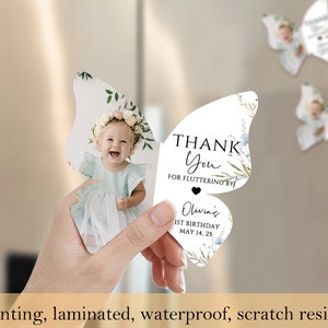 May include: Butterfly-shaped party favour with a photo of a baby girl wearing a floral crown. The favour reads "Thank You for Fluttering By" and "Olivia's 1st Birthday May 14, 25". The image also states "HD printing, laminated, waterproof, scratch resistant".