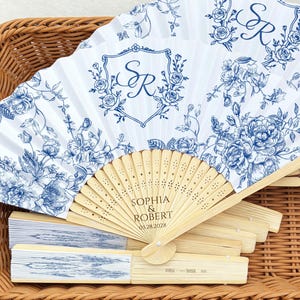 Op de afbeelding: Witte en blauwe vouwventilatoren met bloemenprint en houten handvatten. De ventilatoren hebben een monogram en de namen "Sophia & Robert" met de datum 03.28.2028. De ventilatoren worden in een rieten mand gepresenteerd.