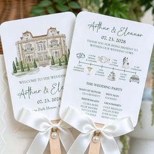 Könnte beinhalten: Zwei weiße Hochzeitsfächer mit Holzgriffen und weißen Satinschleifen mit Perlenakzenten. Ein Fächer zeigt ein Aquarell eines Herrenhauses, der andere einen Zeitplan, die Namen Arthur & Eleanor sowie das Datum 23.07.2028.
