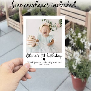 May include: A white card featuring a photograph of a baby girl with a floral crown and a cake. The card says "Olivia's 1st birthday" and "Thank you for celebrating with me! May 14, 2026." The words "free envelopes included" are written at the top.