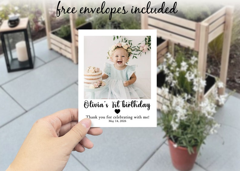 May include: A white card featuring a photograph of a baby girl with a floral crown and a cake. The card says "Olivia's 1st birthday" and "Thank you for celebrating with me! May 14, 2026." The words "free envelopes included" are written at the top.