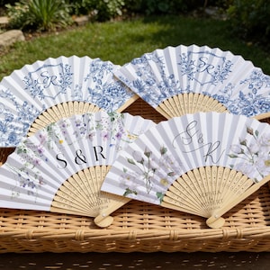 May include: Four decorative hand fans with floral and monogram designs. Two fans feature blue floral patterns on a white background, while the other two have floral and striped designs with the initials "S & R" and "S & K". The fans are made of paper and wood.