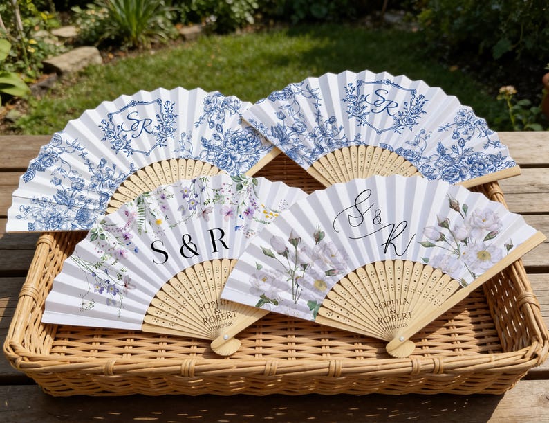May include: Four decorative hand fans with floral and monogram designs. Two fans feature blue floral patterns on a white background, while the other two have floral and striped designs with the initials "S & R" and "S & K". The fans are made of paper and wood.