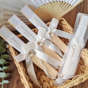 May include: A collection of personalized wedding fans in a woven basket. Each fan features a white paper band with the names "EVELYN & NOAH" and the date "05.28.2028". The fans are adorned with white ribbons and pearl accents.