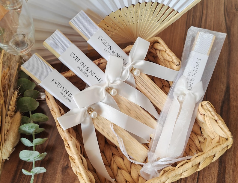 May include: A collection of personalized wedding fans in a woven basket. Each fan features a white paper band with the names "EVELYN & NOAH" and the date "05.28.2028". The fans are adorned with white ribbons and pearl accents.