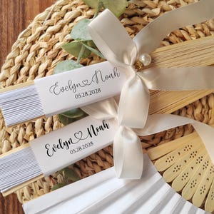 May include: Two white paper fans with the names "Evelyn & Noah" and the date "05.28.2028" printed on them. The fans are tied with ivory satin ribbons and a pearl embellishment. The fans are on a woven placemat.