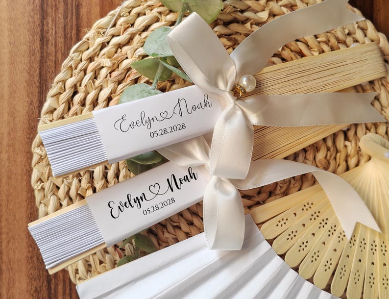 May include: Two white paper fans with the names "Evelyn & Noah" and the date "05.28.2028" printed on them. The fans are tied with ivory satin ribbons and a pearl embellishment. The fans are on a woven placemat.