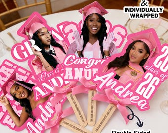 May include: Pink graduation party decorations featuring photos of graduates, the name "Andrea", and the year "26". The decorations include pink bows and wooden sticks with engraved text. The text on the decorations reads "Thanks for celebrating with me!" and "Class of 26".