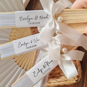 May include: Wooden hand fans with white paper inserts, personalized with the names "Evelyn & Noah" and the date "05.28.2028". Each fan is adorned with a white satin ribbon, a pearl, and a decorative gold accent. Ideal for wedding favors.