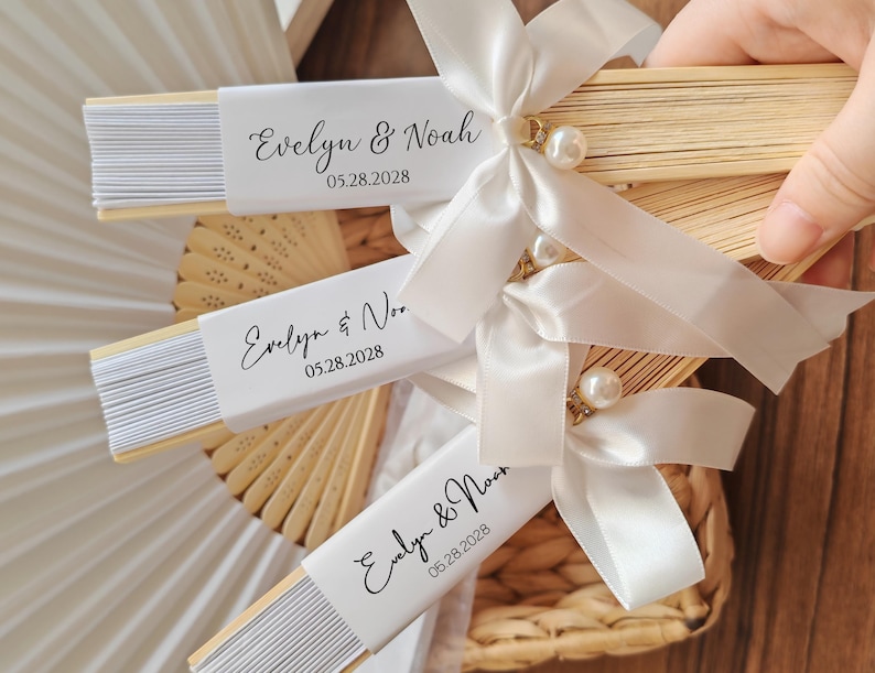 May include: Wooden hand fans with white paper inserts, personalized with the names "Evelyn & Noah" and the date "05.28.2028". Each fan is adorned with a white satin ribbon, a pearl, and a decorative gold accent. Ideal for wedding favors.