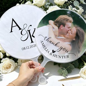 May include: Two round hand fans with clear plastic handles. One fan features the black text A&K and the date 08.12.2028 on a white background. The other fan displays a photograph of a couple with the text thank you AMELIA & HUNTER.