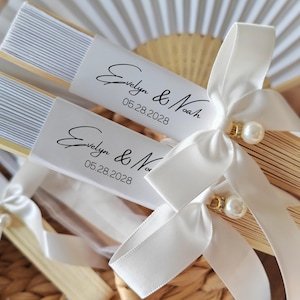 May include: Two personalized paper fans with white ribbon bows and pearl accents. Each fan has a white label with the names "Evelyn & Noah" and the date "05.28.2028" in black script. The fans are made of light-colored wood and white paper.